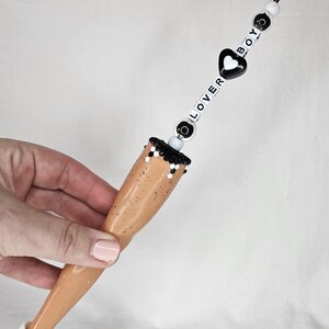 May include: A novelty clip featuring a doll leg design. The leg is tan with a white sneaker. The clip has a beaded chain with the words "LOVER BOY" and a black heart. The clip is held by a hand.