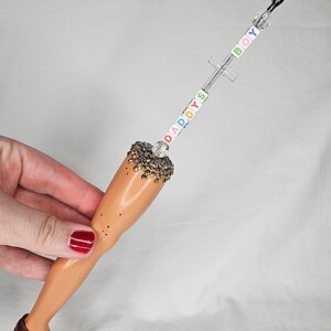 May include: A novelty item featuring a doll leg with a brown shoe adorned with gold studs. The leg is topped with a decorative element and a clip. The words "DADDYS BOY" are spelled out with colorful beads on the clip's stem.