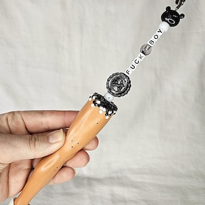 May include: A novelty clip featuring a doll leg with a white sneaker. The clip is adorned with black and white beads, a black bear head, and the text "FUCK BOY". The clip is designed to hold papers or photos.