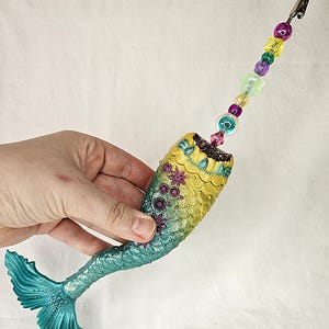 May include: A mermaid tail-shaped clip with a beaded strand. The tail is teal, transitioning to yellow and green with floral accents. The clip is held together by a strand of colorful beads, including purple, yellow, and green.