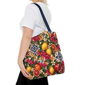 May include: A tote bag featuring a vibrant Mediterranean-inspired print with yellow lemons, red pomegranates, and blood oranges interspersed with pink flowers and blue and white geometric tile patterns, finished with dark navy blue shoulder straps.