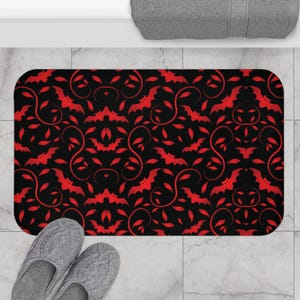 May include: A rectangular bath mat with a repeating pattern of red bats and vines on a black background. The mat is on a tiled floor, with a pair of gray slippers in the foreground and a gray towel in the background.