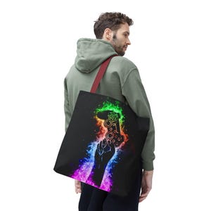 May include: A black tote bag with a colorful design of a hand holding a video game controller. The controller is outlined in green and surrounded by rainbow flames.