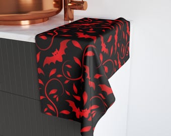 May include: Black hand towel with a repeating pattern of red bats and leaves. The towel is draped over a white countertop, with a copper sink and faucet in the background. The design is suitable for Halloween.