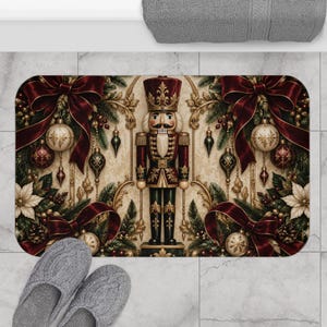 May include: A rectangular bath mat featuring a central nutcracker figure in a red and gold uniform, surrounded by dark red bows, gold ornaments, green pine branches, and white poinsettia flowers on a cream-colored background with gold scrollwork.