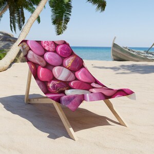 May include: A beach chair featuring a fabric cover with a repeating pattern of smooth, polished stones in various shades of pink, magenta, and white, some with gold-colored veining, supported by a light-colored wooden frame on a sandy beach.