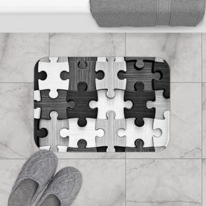May include: A rectangular bath mat featuring a puzzle piece design in shades of black, white, and gray. The mat has a non-slip backing and is designed to absorb water. The mat is placed on a tiled floor.
