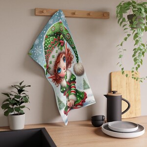 May include: A Christmas-themed kitchen towel featuring a whimsical elf illustration. The elf has red curly hair, blue eyes, and wears a green and red outfit with a matching hat. The background is a snowy scene with snowflakes and Christmas trees.