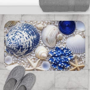 May include: A rectangular bath mat featuring a beach theme. The mat is adorned with seashells, starfish, and blue coral on a bed of white pebbles. A large blue and white seashell is a focal point. Gray slippers are at the bottom.