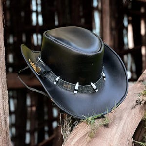 May include: A black leather cowboy hat with a wide brim and a tall crown. The hat features a decorative band with white accents and a chin strap. A small gold-colored emblem is visible on the side. The hat is resting on a wooden beam.