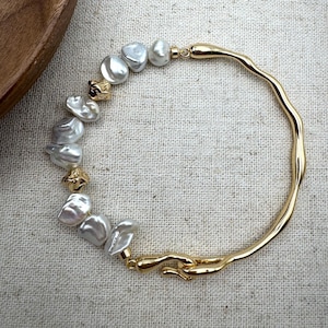 Puede incluir: Un brazalete dorado con un diseño ondulado, adornado con una fila de perlas blancas irregulares.