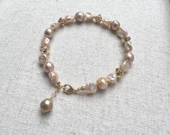 Peut inclure: Un bracelet délicat orné de perles de couleur claire de forme irrégulière, dans des tons de rose, de crème et d'or. Les perles sont enfilées avec des accents métalliques dorés et un fermoir assorti. Un seul charme de perle pend du fermoir.