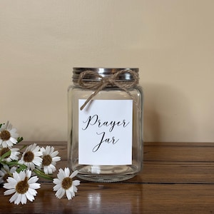 May include: A clear glass jar with a silver lid and a brown twine bow. The jar has a white label with the text "Prayer Jar" in black cursive writing.