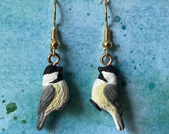 May include: A pair of gold dangle earrings featuring a small, detailed bird charm. The bird is painted in shades of black, white, and gray, resembling a chickadee.