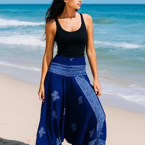May include: A person wearing a black vest top and blue harem trousers with a white floral pattern. The trousers have an elasticated waistband and are loose-fitting. The background is a beach with blue sea and sand.