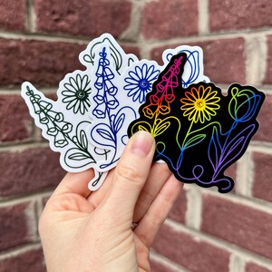 K&ouml;nnte beinhalten: Eine Hand h&auml;lt eine Sammlung von Blumenaufklebern. Die Aufkleber zeigen Strichzeichnungen verschiedener Blumen, darunter G&auml;nsebl&uuml;mchen und Fingerhut. Ein Aufkleber ist schwarz mit einem Regenbogenverlauf, andere sind wei&szlig; mit farbigen Umrissen.