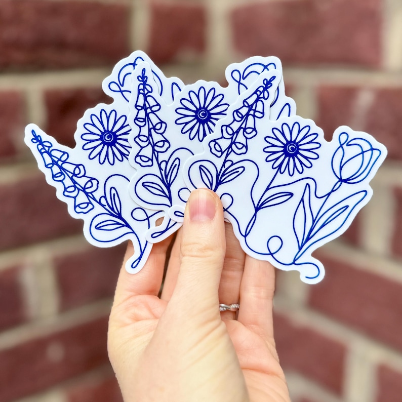 K&ouml;nnte beinhalten: Eine Hand h&auml;lt einen Stapel wei&szlig;er Aufkleber mit blauen floralen Linienzeichnungen. Die Aufkleber zeigen verschiedene Blumen, darunter G&auml;nsebl&uuml;mchen, Fingerhut und Tulpen. Der Hintergrund ist eine rote Backsteinmauer.