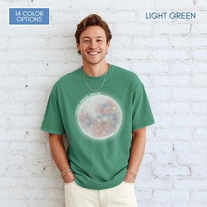 May include: Light green t-shirt with a graphic of a colorful moon and the word "AMAZE" repeated around the moon. The shirt also has the text "14 COLOR OPTIONS" in a blue rectangle and "LIGHT GREEN" in a green rectangle.