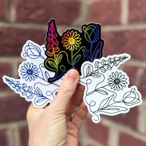 May include: A hand holds several floral stickers. The stickers feature line art designs of various flowers, including daisies, tulips, and foxgloves. Some stickers have a black background with rainbow-colored flowers, while others are white with blue or black outlines.
