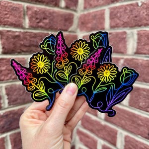K&ouml;nnte beinhalten: Eine Hand h&auml;lt mehrere schwarze Aufkleber mit farbenfrohen floralen Designs. Die Aufkleber zeigen Umrisse von Blumen und Bl&auml;ttern in einem Regenbogenverlauf auf schwarzem Hintergrund. Die Blumen umfassen G&auml;nsebl&uuml;mchen, Fingerhut und Tulpen.
