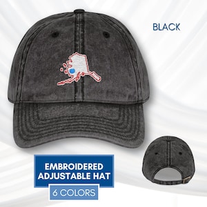 Könnte beinhalten: Eine schwarze Baseballkappe aus verwaschenem Denim mit einer weißen Stickerei, die den Umriss des Bundesstaates Alaska mit einem kleinen blauen Punkt zeigt. Die Kappe hat einen gebogenen Schirm, sechs Paneele mit Ösen und einen verstellbaren Metallriemen.