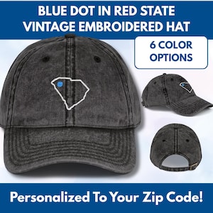 Può includere: Cappello vintage ricamato grigio scuro con un contorno bianco di uno stato e un punto blu. Il testo recita "BLUE DOT IN RED STATE" e "6 COLOR OPTIONS". L'immagine mostra anche "Personalized To Your Zip Code!"
