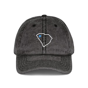 Puede incluir: Gorra de béisbol negra con visera curvada. La gorra presenta un contorno blanco del estado de Carolina del Sur con un punto azul, bordado en la parte delantera. El sombrero tiene un aspecto vintage.
