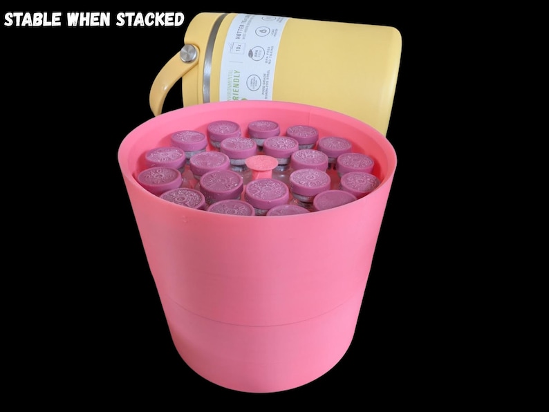 May include: A pink cylindrical container filled with multiple small glass vials topped with purple caps. Behind the container sits a yellow insulated bottle with a handle. The text STABLE WHEN STACKED is visible in the upper left corner.