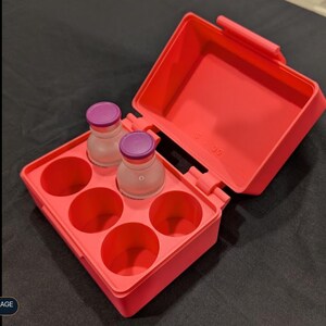 May include: A bright coral-colored rectangular storage case with an open hinged lid, featuring six circular compartments. Two small clear glass vials with purple caps are placed inside two of the slots, while four slots remain empty.