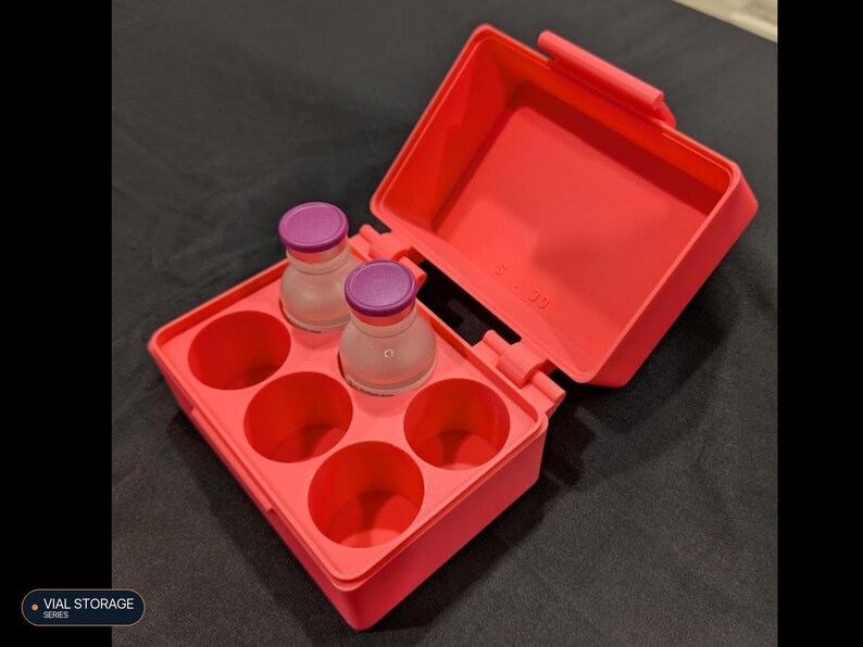 May include: A bright coral-colored rectangular storage case with an open hinged lid, featuring six circular compartments. Two small clear glass vials with purple caps are placed inside two of the slots, while four slots remain empty.