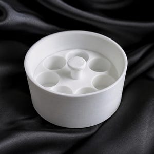 May include: A white circular organizer tray featuring a central handle and seven evenly spaced cylindrical compartments arranged in a ring. The object has a smooth, matte finish and a uniform structure designed for holding small items.