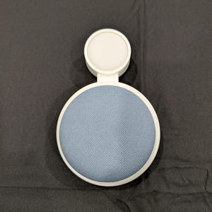 Puede incluir: Un soporte de dispositivo de pl&aacute;stico blanco con una secci&oacute;n de altavoz circular cubierta de tela azul y un accesorio de base de carga blanco m&aacute;s peque&ntilde;o. El texto DEVICE MOUNTS SERIES es visible en la esquina superior derecha.