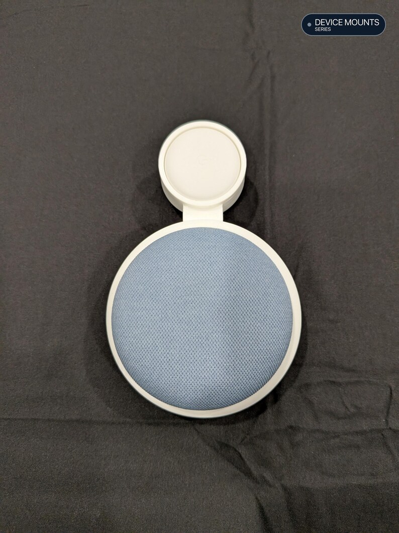 Puede incluir: Un soporte de dispositivo de pl&aacute;stico blanco con una secci&oacute;n de altavoz circular cubierta de tela azul y un accesorio de base de carga blanco m&aacute;s peque&ntilde;o. El texto DEVICE MOUNTS SERIES es visible en la esquina superior derecha.