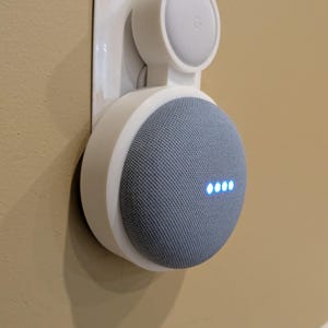 Puede incluir: Un soporte de pared de pl&aacute;stico blanco que sostiene un altavoz inteligente circular de color gris. El altavoz presenta un frente cubierto de tela con cuatro luces indicadoras azules iluminadas. El soporte est&aacute; fijado a un enchufe de pared.