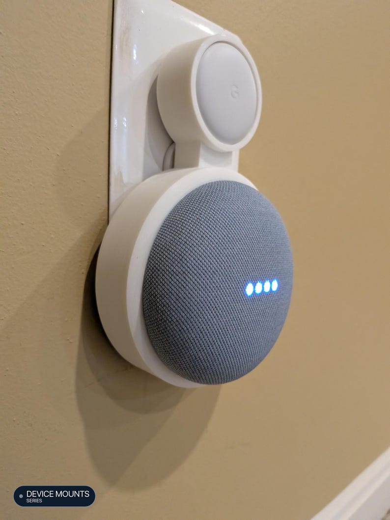 Puede incluir: Un soporte de pared de pl&aacute;stico blanco que sostiene un altavoz inteligente circular de color gris. El altavoz presenta un frente cubierto de tela con cuatro luces indicadoras azules iluminadas. El soporte est&aacute; fijado a un enchufe de pared.
