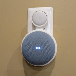 May include: A white wall mount for a Google Home Mini smart speaker. The speaker is a light blue color with a fabric covering and two blue lights on the front. The speaker is mounted on a white plastic wall mount.