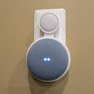 Puede incluir: Un soporte de pared de pl&aacute;stico blanco que sostiene un altavoz inteligente circular gris con una cubierta de tela y dos luces indicadoras azules iluminadas. El soporte tiene una secci&oacute;n superior circular con el logo de Google.