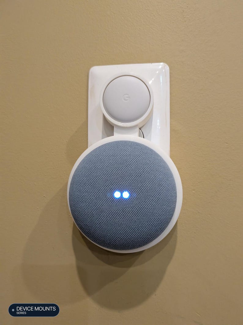 Puede incluir: Un soporte de pared de pl&aacute;stico blanco que sostiene un altavoz inteligente circular gris con una cubierta de tela y dos luces indicadoras azules iluminadas. El soporte tiene una secci&oacute;n superior circular con el logo de Google.