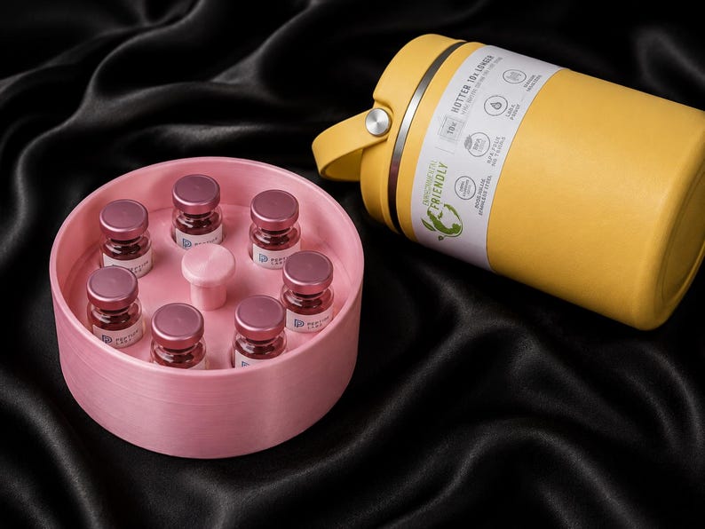 May include: A yellow insulated stainless steel bottle with a handle and a label reading HOTTER 10X LONGER, alongside a round pink container holding seven small glass vials with metallic pink caps arranged in a circular pattern.