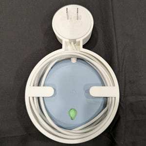 Puede incluir: Un soporte de pared de pl&aacute;stico blanco para altavoz inteligente, con un marco circular para organizar cables y un enchufe integrado. El soporte sostiene un dispositivo circular azul claro con una peque&ntilde;a luz indicadora verde al frente.