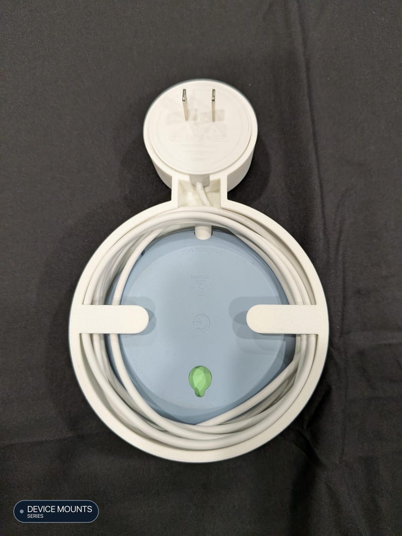 Puede incluir: Un soporte de pared de pl&aacute;stico blanco para altavoz inteligente, con un marco circular para organizar cables y un enchufe integrado. El soporte sostiene un dispositivo circular azul claro con una peque&ntilde;a luz indicadora verde al frente.