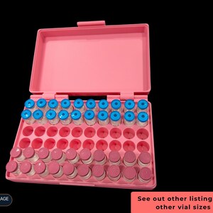 Op de afbeelding: Een roze plastic opbergdoos met een open deksel, met meerdere rijen kleine glazen flesjes. De bovenste rij bevat flesjes met felblauwe doppen, terwijl de onderste rijen flesjes met roze doppen bevatten. De doos heeft lege ronde uitsparingen.
