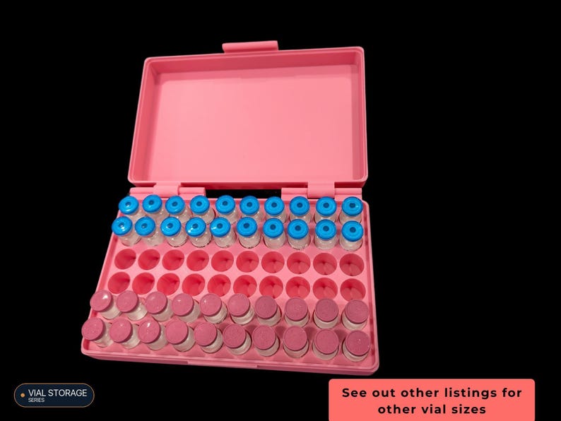 Op de afbeelding: Een roze plastic opbergdoos met een open deksel, met meerdere rijen kleine glazen flesjes. De bovenste rij bevat flesjes met felblauwe doppen, terwijl de onderste rijen flesjes met roze doppen bevatten. De doos heeft lege ronde uitsparingen.