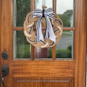 May include: A nautical-themed wreath made of thick, braided rope in shades of tan, white, and navy blue. A large bow with navy and white striped ribbon and white streamers adorns the top. The wreath hangs on a wooden door with glass panels.