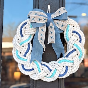May include: A nautical-themed wreath made of thick, braided white rope, accented with sections of light blue and navy blue rope. A large blue bow with starfish patterns adorns the top. This decorative wreath is perfect for coastal decor.
