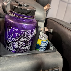 May include: A purple water bottle with a black lid and a white topographical map design. The bottle is in a car's cup holder next to a can of soda.