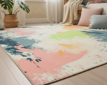 May include: A rectangular area rug with a cream-colored background and colorful paint splotches in pink, blue, yellow, and green. The rug is placed on a light-colored wooden floor, with a potted plant and window in the background.