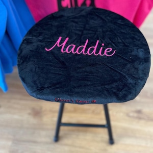 May include: Black velvet stool with the name "Maddie" embroidered in pink script. The stool has a black metal frame and a round seat. The text "Glam's Closet" is embroidered in red on the seat.