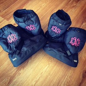May include: Four pairs of navy blue dance booties with pink embroidered initials. The booties are quilted and have a velcro strap. The brand name "BLOCK" is printed in white on the side of each bootie.