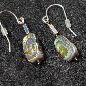 May include: A pair of dangle earrings featuring iridescent abalone shell beads with a rectangular shape. Each earring includes a silver-toned metal hook, a small metallic spacer bead, and a clear silicone earring back for secure placement.